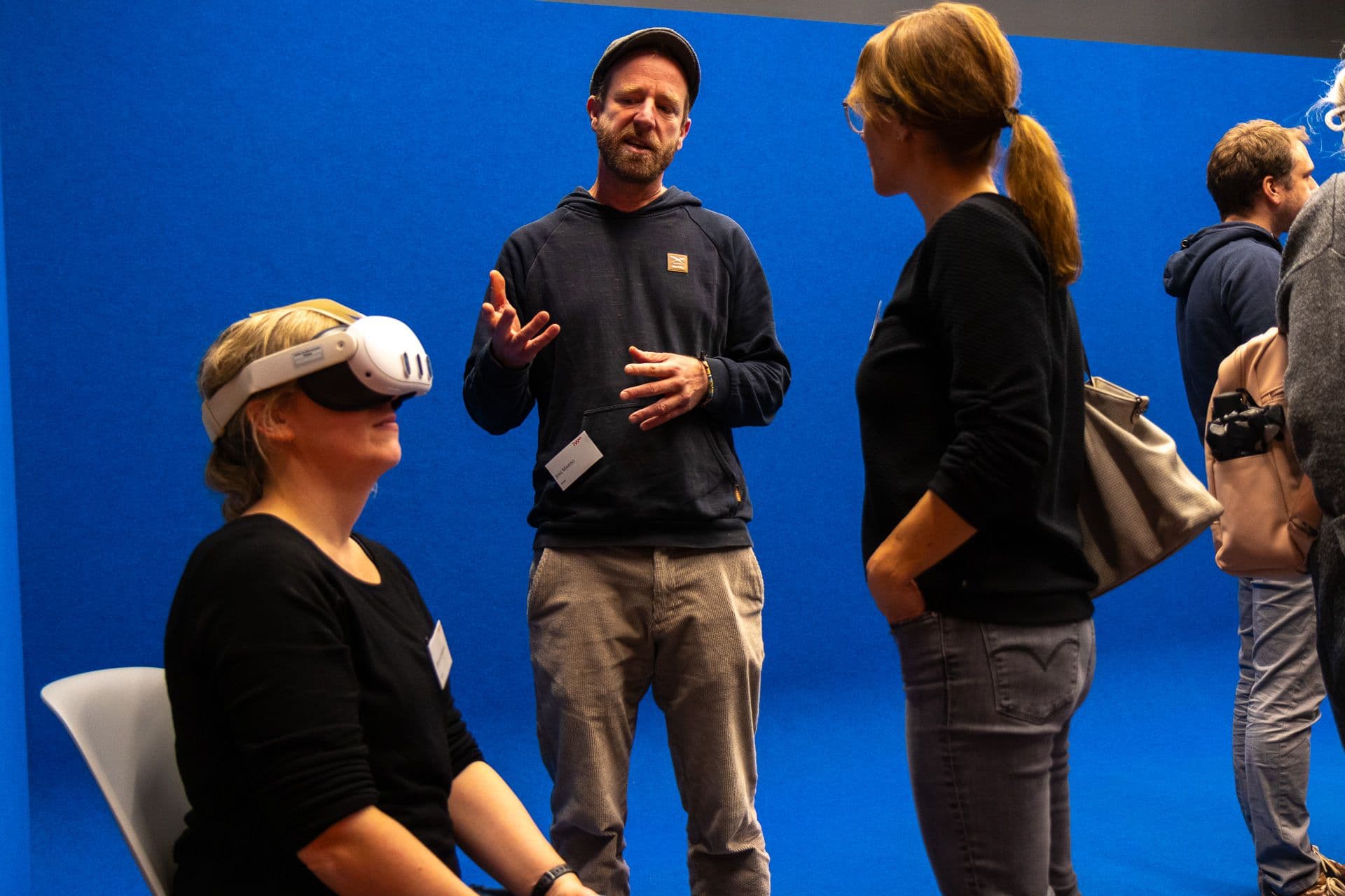 IWM alumni chatting in front of a blue background. Person with VR glasses in the foreground.