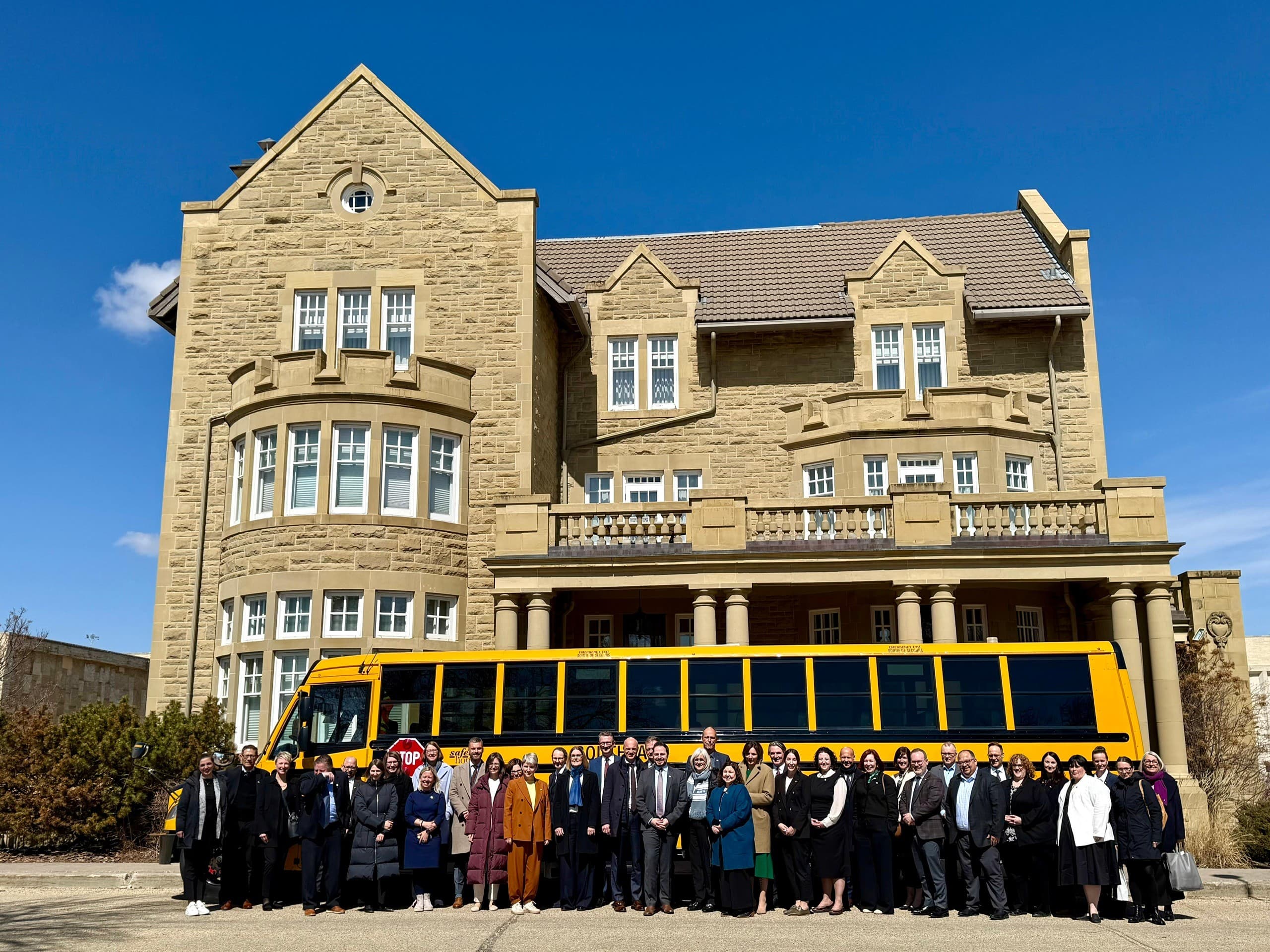 A German delegation stands in front of the Ministry of Education of the Canadian province of Alberta. The group poses in front of a historic sandstone building, with a yellow school bus visible behind them. The sky is clear and blue.